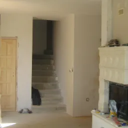 Wnętrze w trakcie remontu: widoczne schody betonowe, drzwi zabezpieczone folią malarską, niedokończony kominek z białych kafli i ściany przygotowane do malowania.